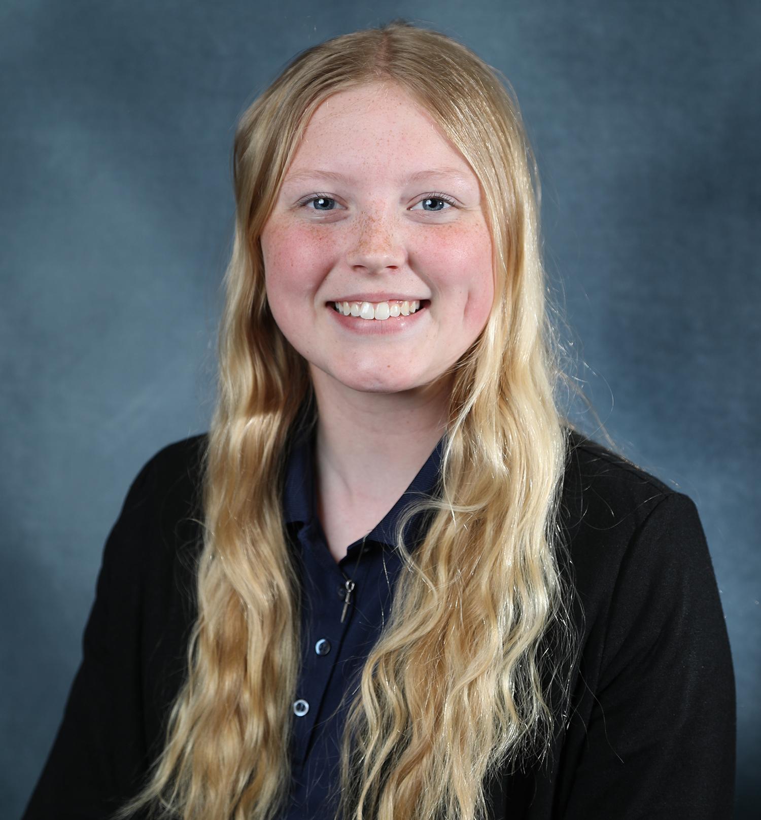 This is a headshot photograph of scholarship winner Kaylie McCutcheon. She is standing in front of a gray backdrop and wearing a black shirt. She is smiling.