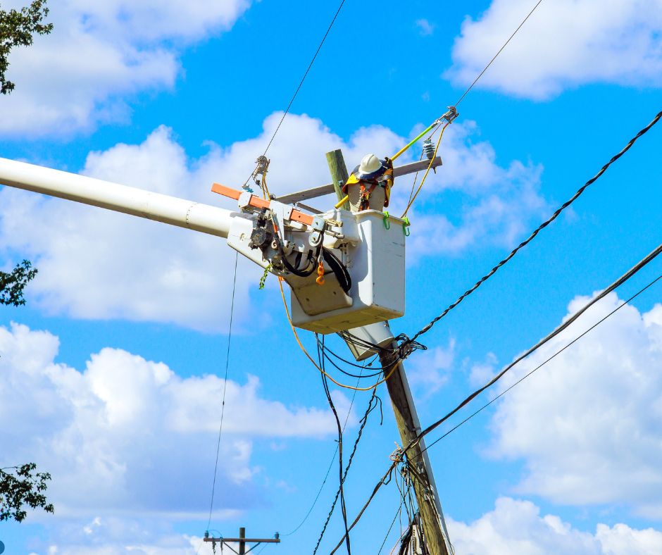 A lineman in a bucket truck repairs a power line against a bright blue sky and white puffy clouds.